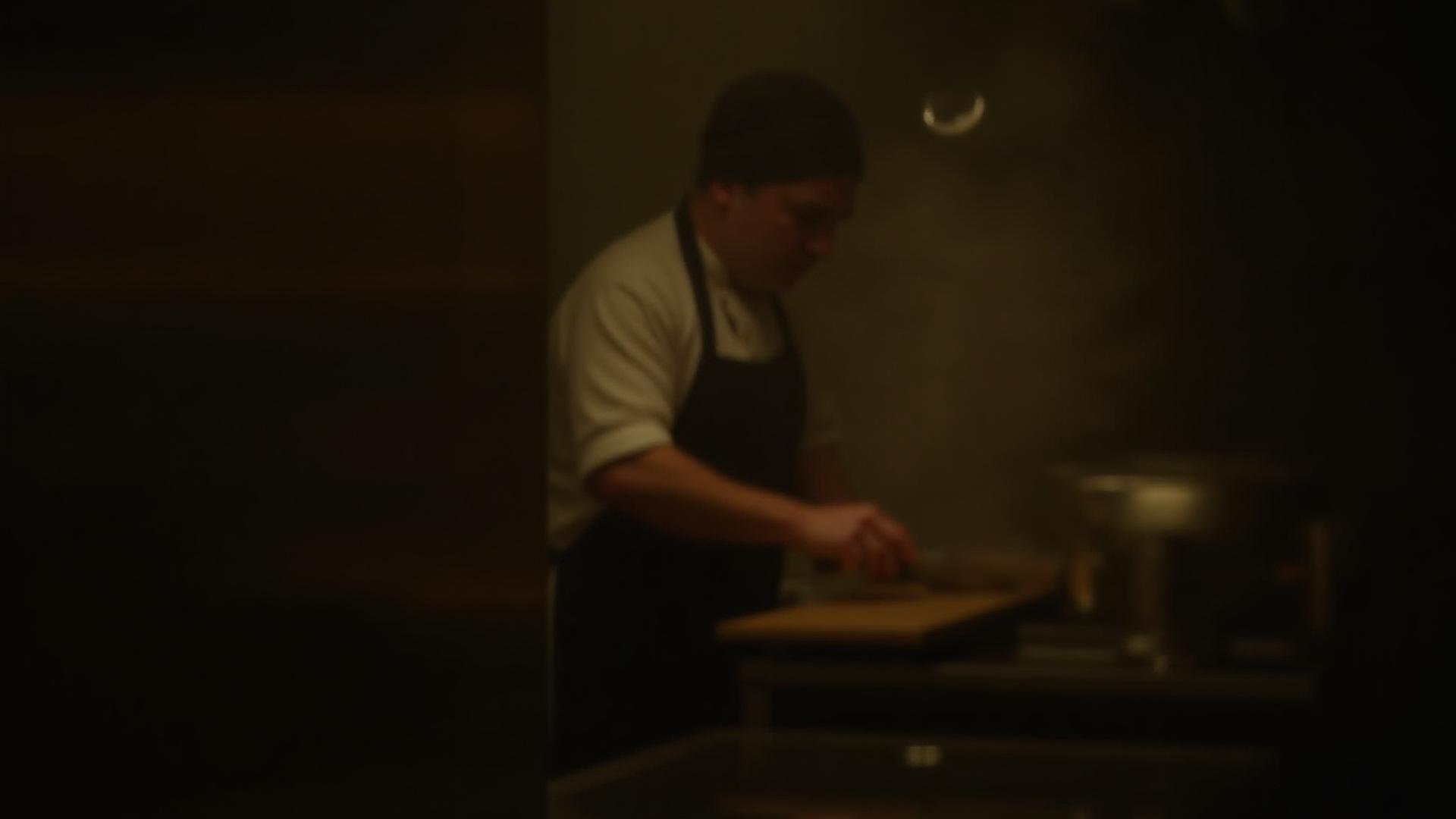 A dimly lit, atmospheric professional kitchen with a chef working in the background, creating a sense of focus and craftsmanship.